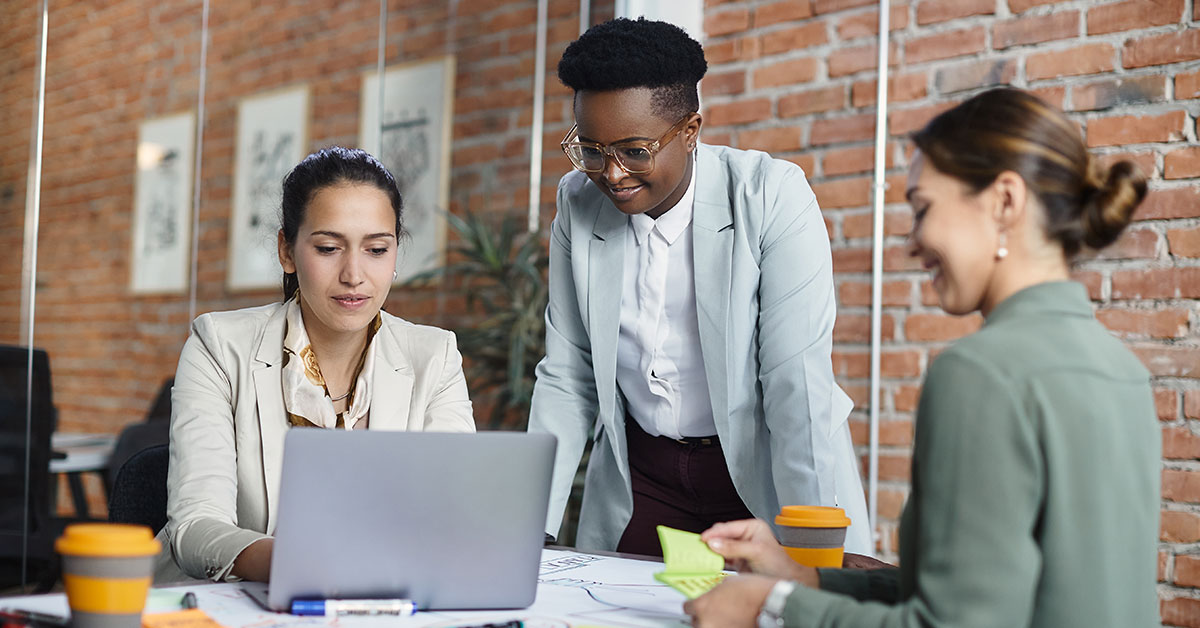 three women working at a desk while two are on the computer, and another one is writing