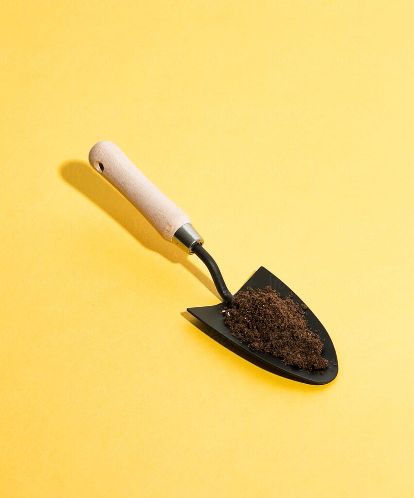 A small garden trowel with a wooden handle and black metal blade, cradling a mound of soil, rests on a bright yellow background, symbolizing the tools essential for cultivating a feedback-rich culture.