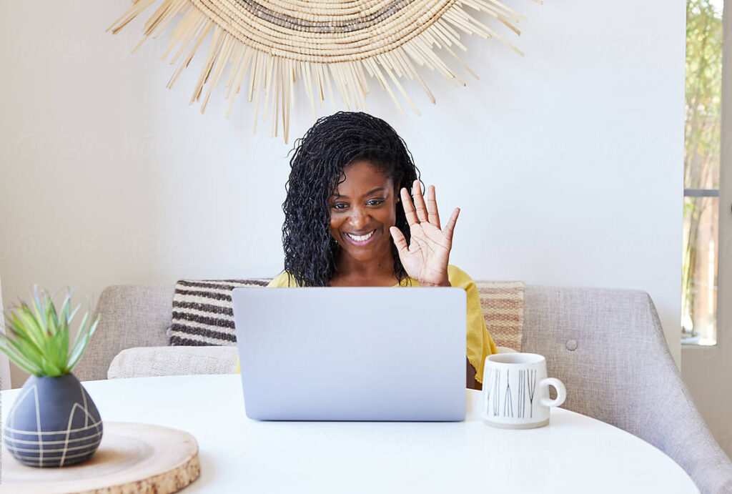 A woman with curly hair sits at a round table, smiling and waving at a laptop. Shes wearing a yellow shirt, embodying her journey in creating your path to professional growth. A coffee mug and a small plant adorn the table, while a decorative piece graces the wall behind her.