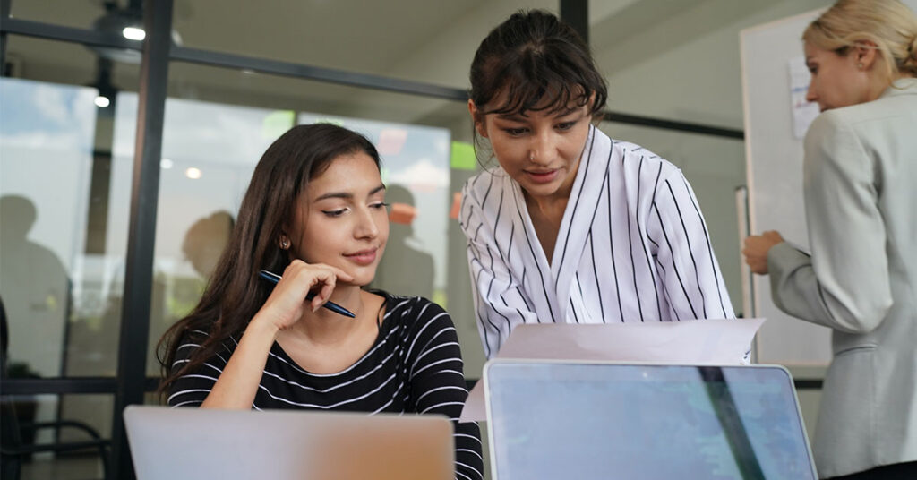 two women working on the computer while talking and another woman in the background of the photo with a white board