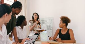 A group of women of color sit around a table and look at another woman in a white dress who is presenting in front of a whiteboard