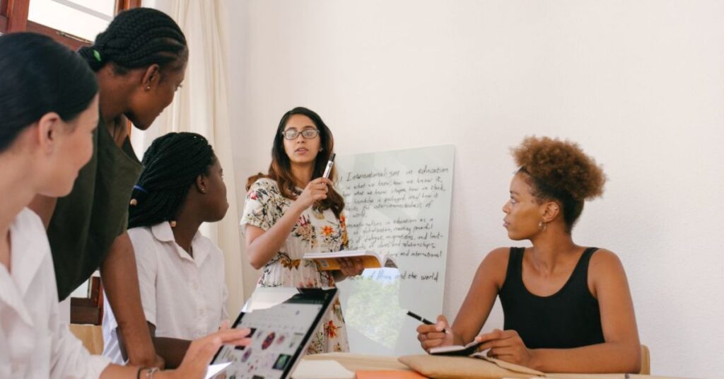 An Asian woman with a marker stands in front of a whiteboard while talking and collaborating with a team seated & standing around a table