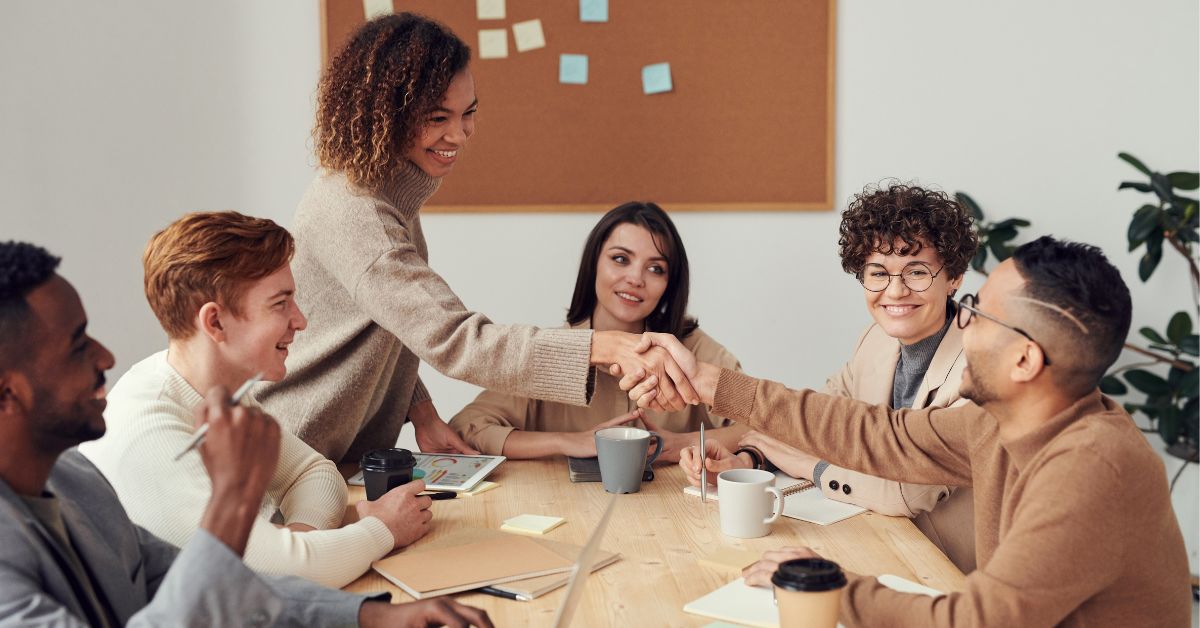 A group of young professionals of color seated around a table are greeting a new female teammate who is standing and shaking hands with a man in glasses with dark hair.