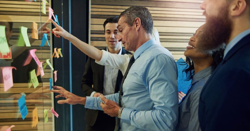 A diverse group of people stand in front of a clear board with colorful sticky notes on it, brainstorming and working together