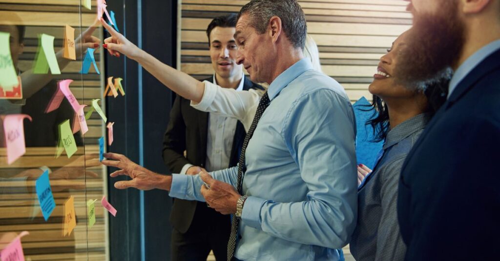 A white man in a blue shirt talks to colleagues as the place colorful sticky notes on a clear whiteboard