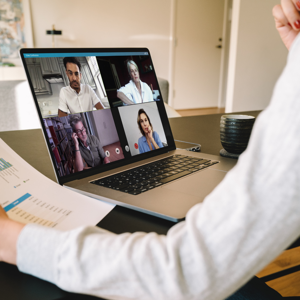 A computer on a desk shows a screen with four people in a meeting with the person sitting at the desk who holds a piece of paper.