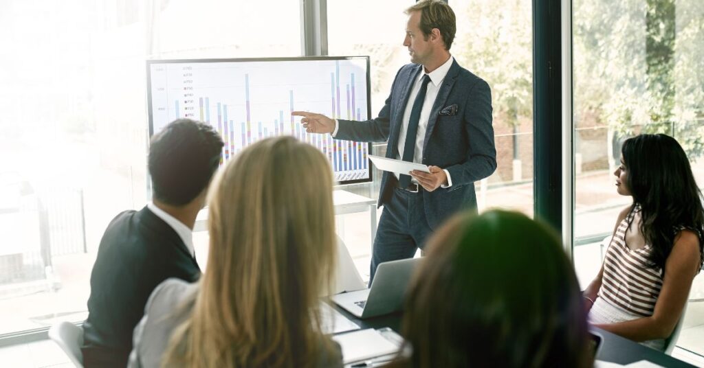 A white man in a suit stands in front of a screen and presents a graph to people seated at a table