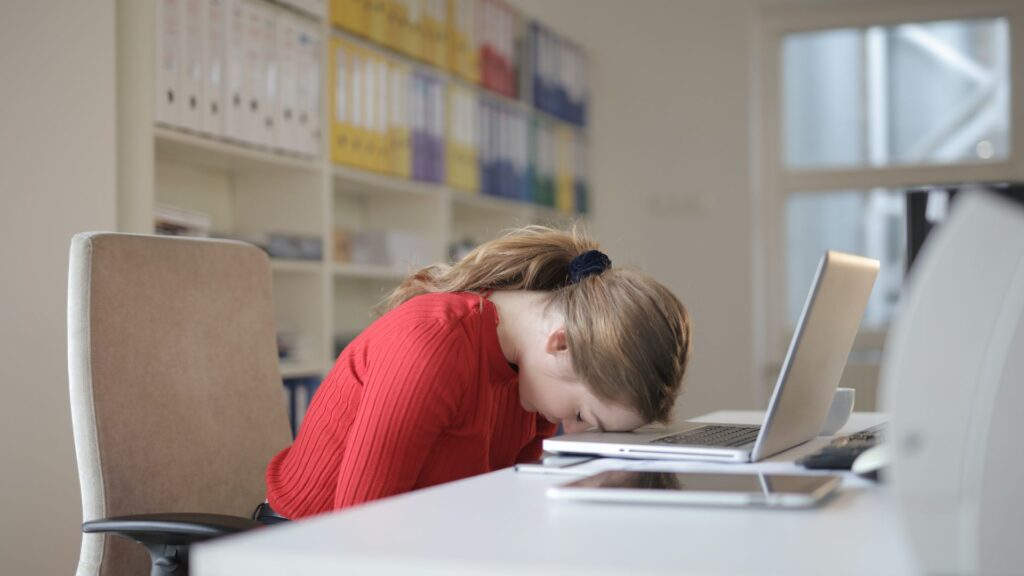 A woman at work wearing a red sweater puts her head on her computer in frustration