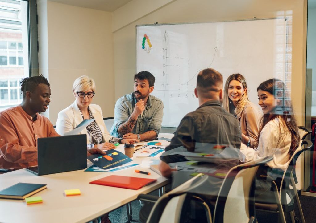 A group of multi-racial & multi age people sit around a table in a conference room, talking animatedly to one another