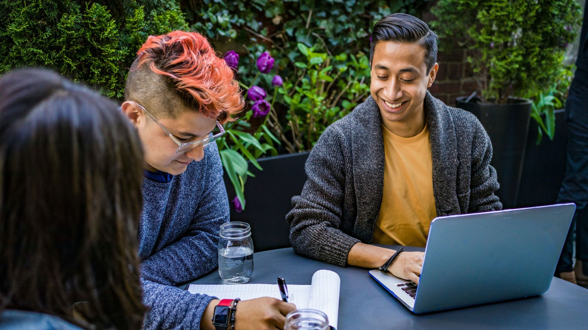 A group of three people sit at a table outside, one brown man with a computer, one white person with a short, pink haircut, and a third person with dark long hair. They're collaborating on work.