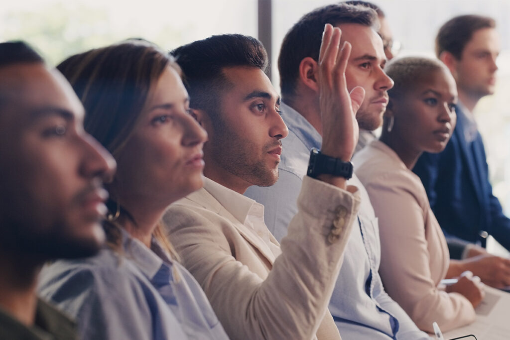 leadership learning event's audience, featuring the question of a business man with hand up at a seminar, workshop or training. Diversity men and women crowd at a presentation for learning, knowledge and faq discussion